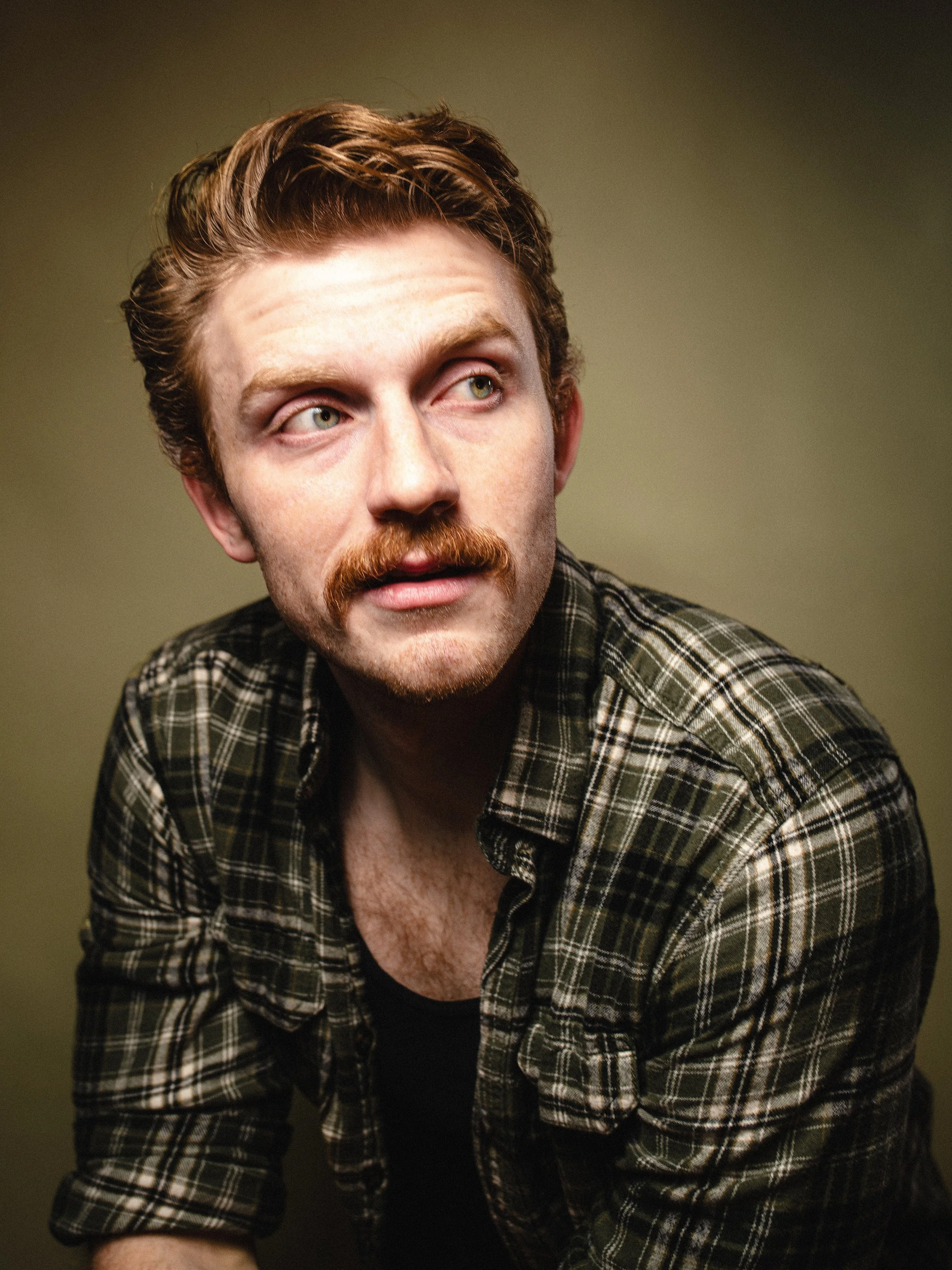 A young man with reddish hair, a mustache, and a beard wearing a plaid shirt, looking to the side.