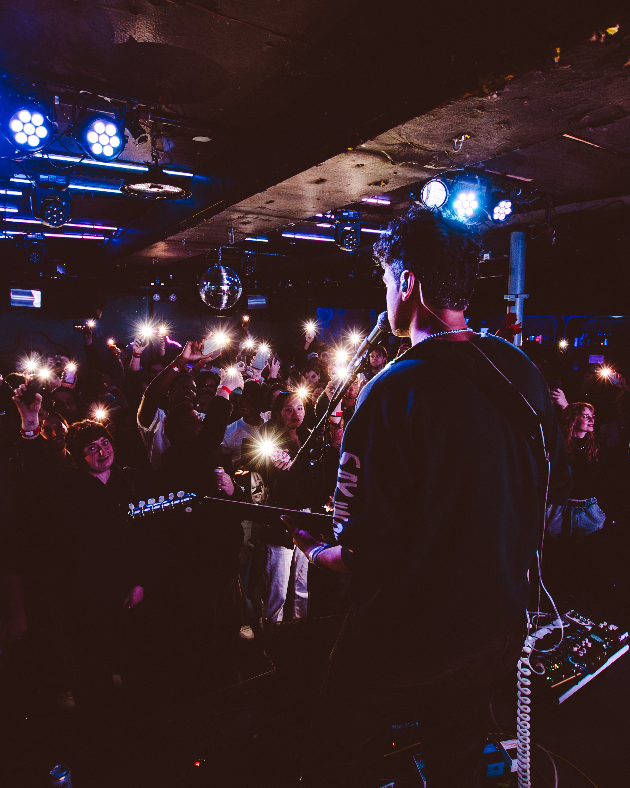 Musician on stage performing at a concert. The audience is holding up lights, creating a bright, lively atmosphere with dark surroundings and colorful stage lighting.