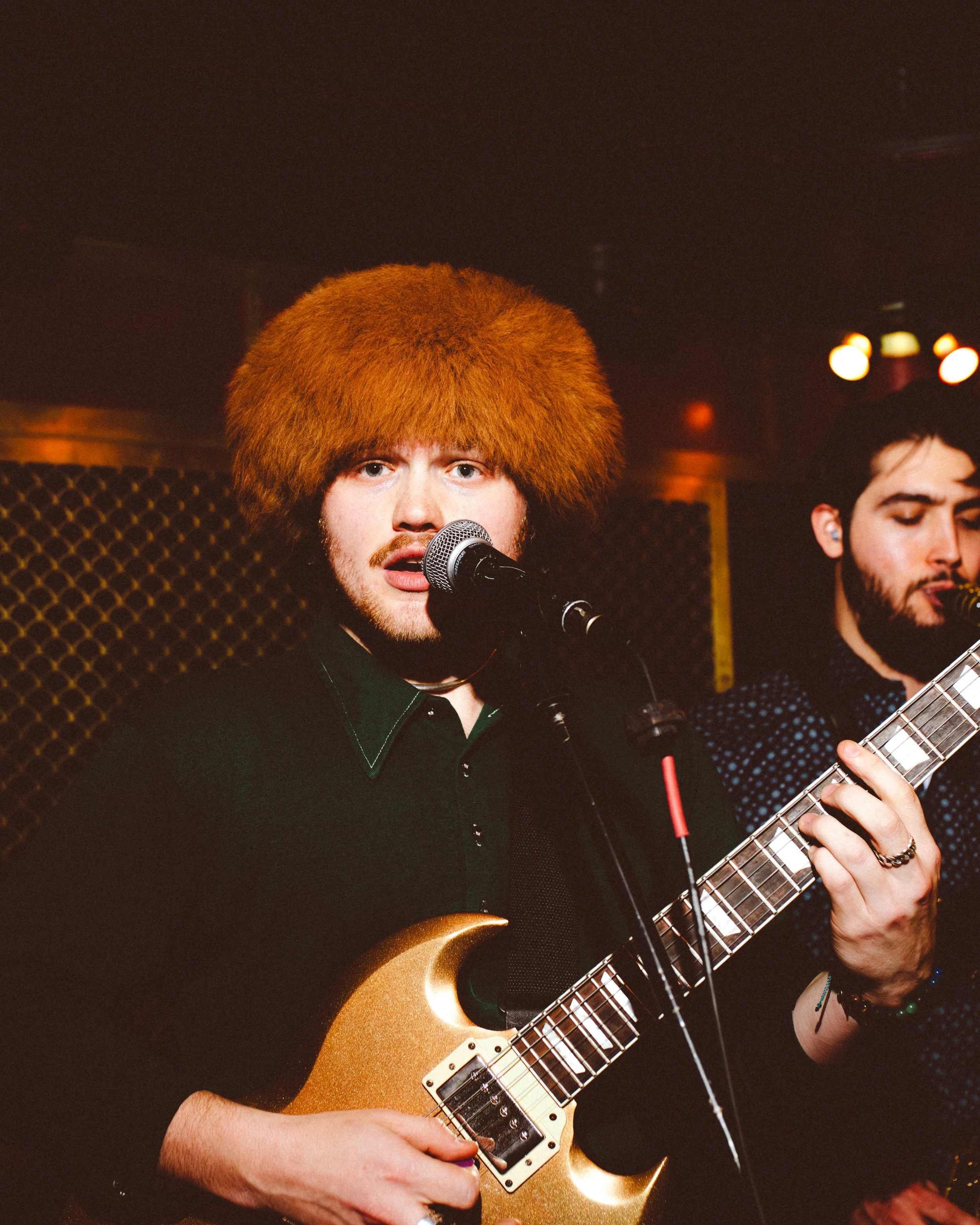 A man with a large orange furry hat playing an electric guitar and singing into a microphone on stage.