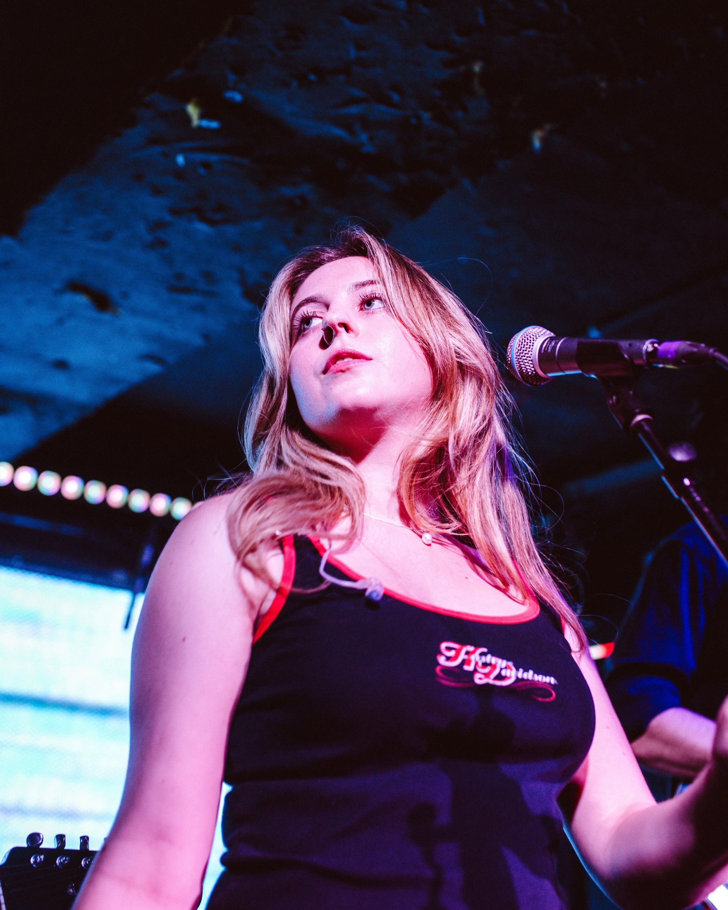 A young woman singing into a microphone on stage, with colorful stage lighting and a dark ceiling above.