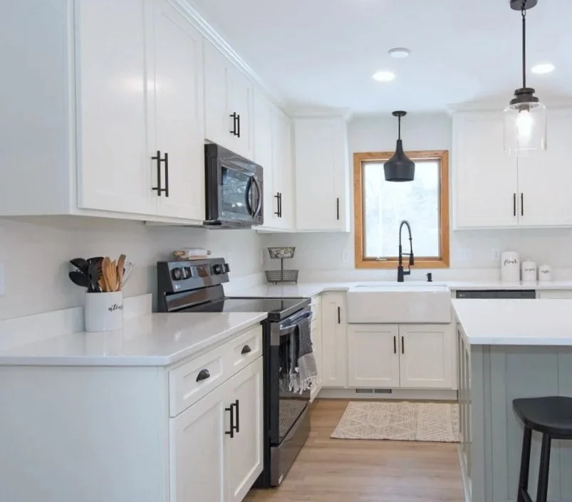 Modern white kitchen with black hardware, black stove and microwave, white countertops, farmhouse sink, window with wooden frame, and simple decor.