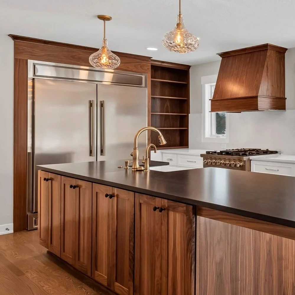 MNBR modern kitchen remodel with wooden cabinets, a black countertop island with a gold faucet, stainless steel appliances including a double-door refrigerator, a range with a wooden hood, open wooden shelves, and pendant lights.