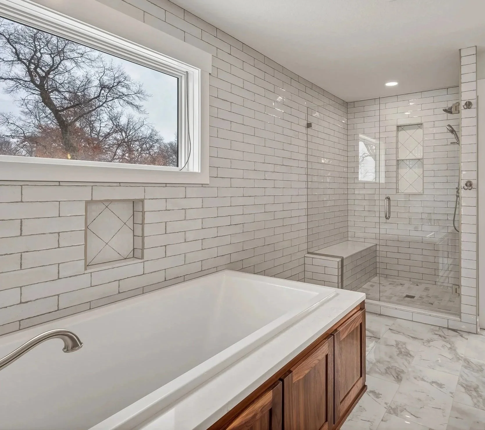 MNBR modern bathroom renovation with white subway tile walls, a bathtub with a wood vanity, a frameless glass shower with built-in niche, marble floor, and a window showing tree branches outside.