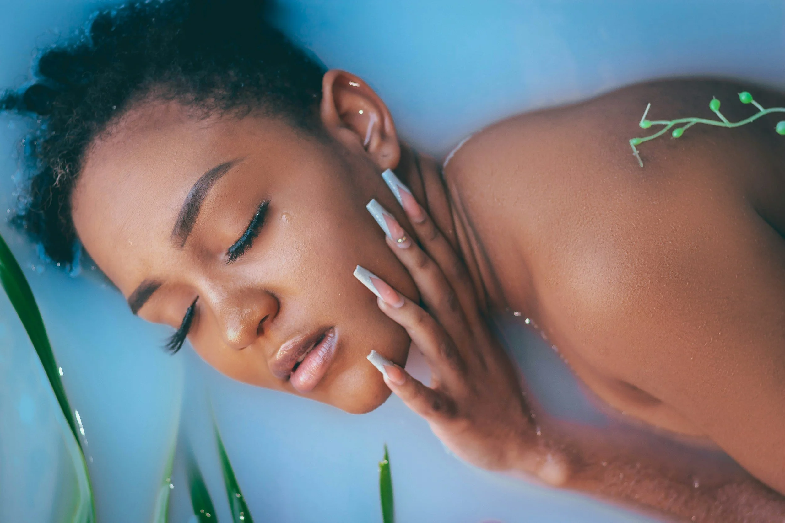 Close-up of a woman with dark skin and short curly hair lying in a bath with water and green plants, her eyes closed and touching her face with one hand.