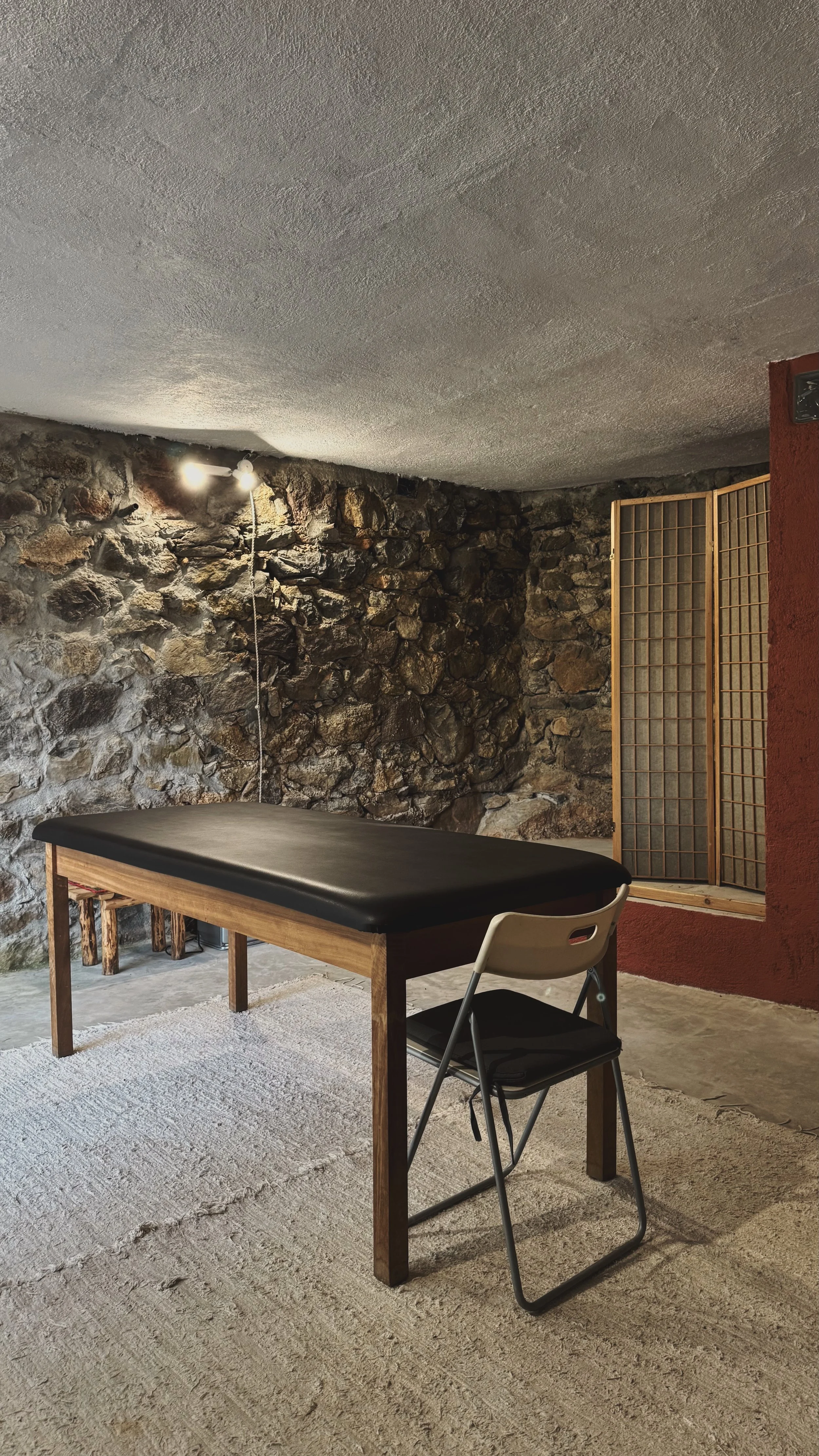A room with an exposed stone wall, a rectangular table with a black top, two chairs (one white and one black), a textured ceiling, and a wooden room divider with a grid pattern. The room has a beige carpet and a ceiling light fixture.