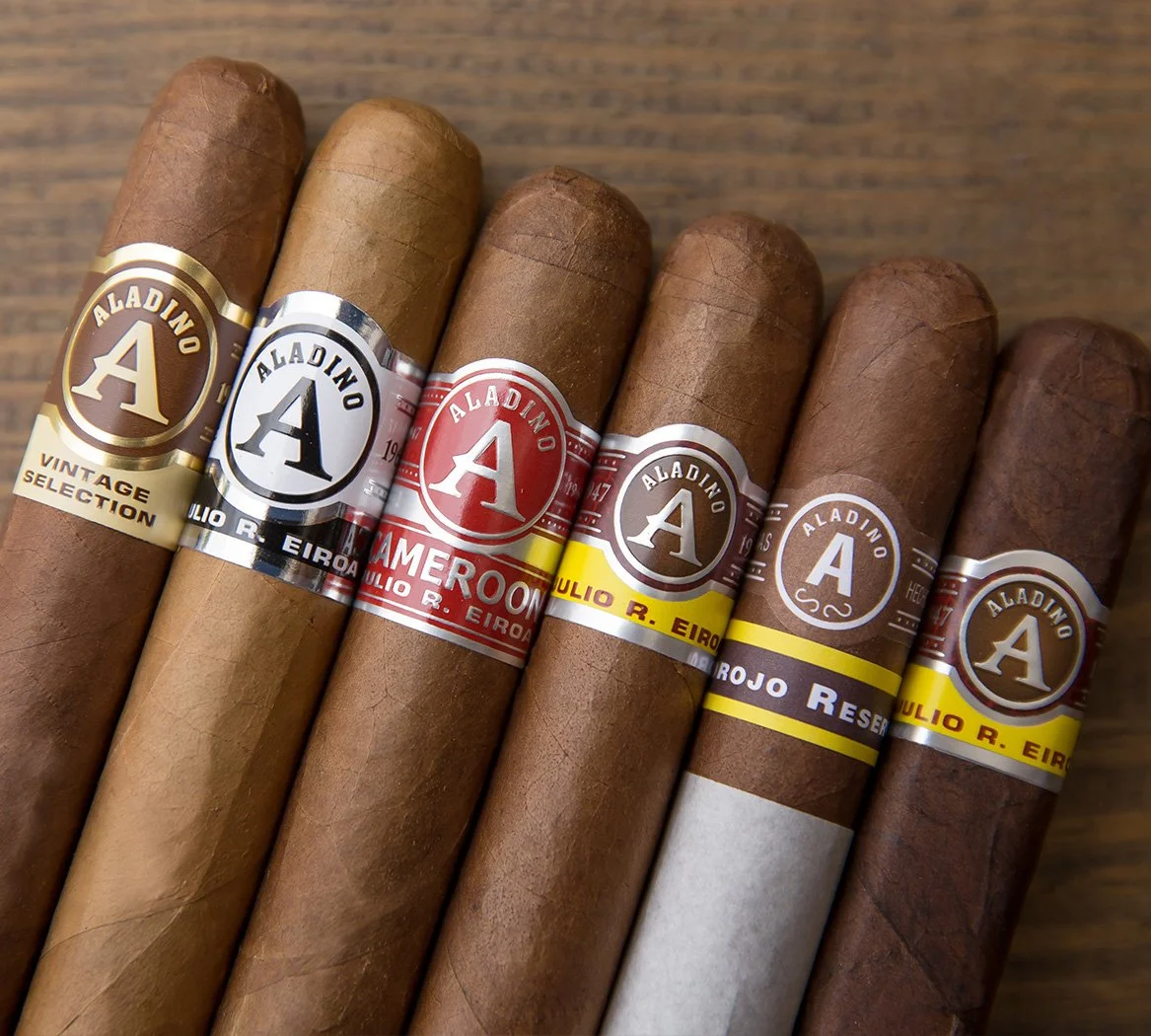 Six cigars lined up on a wooden surface, each with a different colored band and label.
