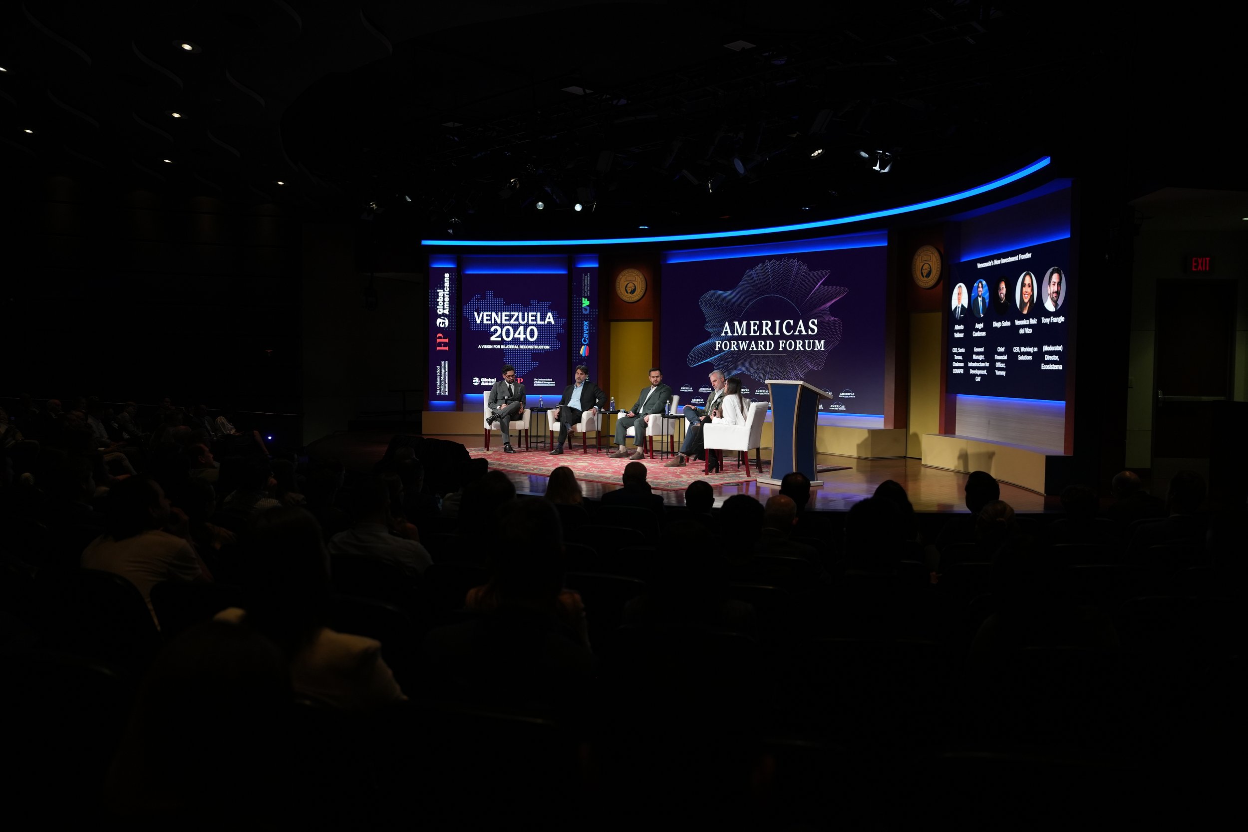 A stage at the front of an auditorium. Five people are on stage, seated in front a background of 5 screens featuring event-branded designs.