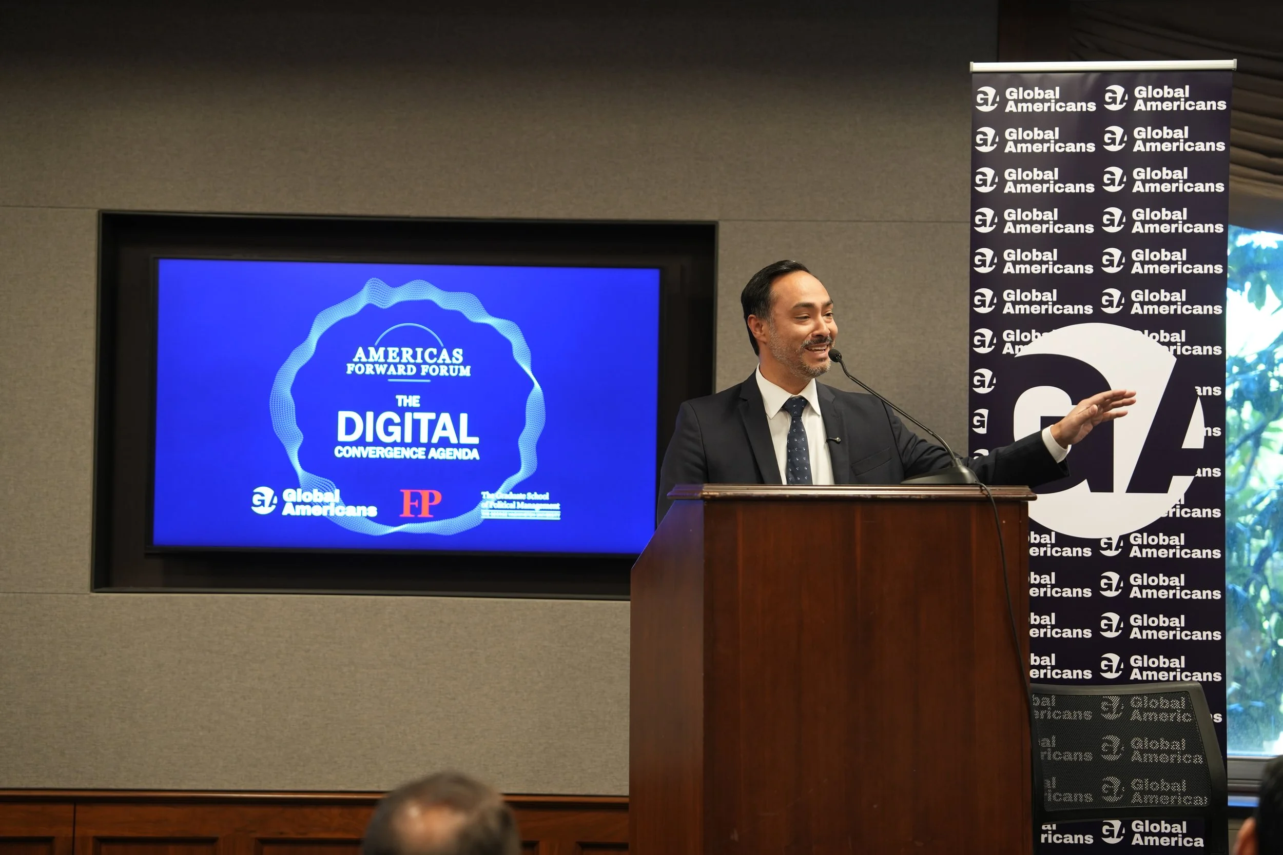 Rep. Joaquin Castro speaking at a lecter, with an event branded banner and screen to his sides.