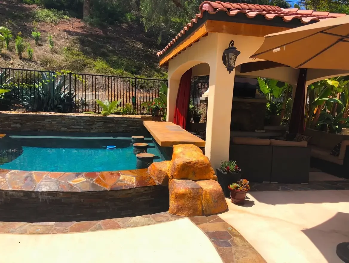 A backyard pool area with blue water, stone edging, and a small seating area with a beige umbrella. There is a shaded outdoor lounge with cushions under a pergola, potted plants, and a view of a hillside with trees and plants in the background.