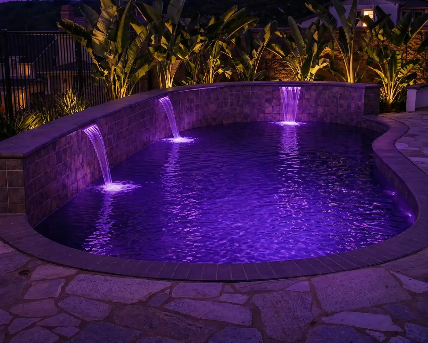 A backyard pool illuminated with purple lights at night, featuring three small waterfalls on a tiled wall, surrounded by tropical plants and stone-paved deck.