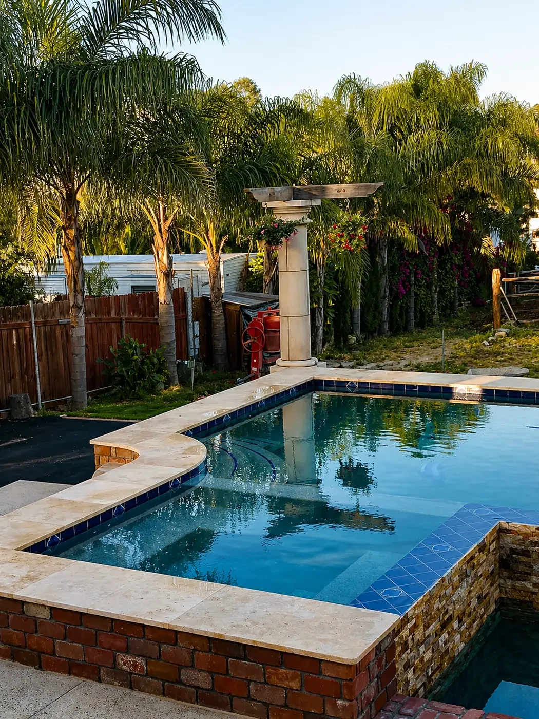 A backyard swimming pool with a limestone deck, surrounded by palm trees and a wooden fence, during sunset.