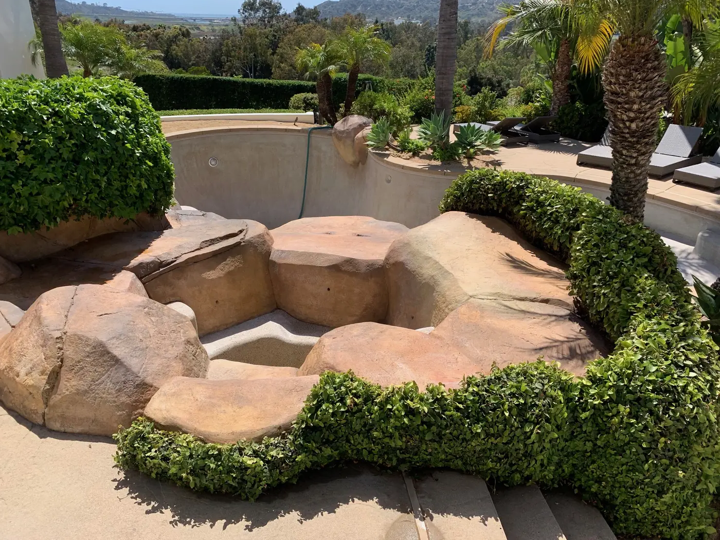 Empty outdoor hot tub surrounded by rocks and green bushes, with palm trees and lounge chairs in the background, in a sunny garden setting.