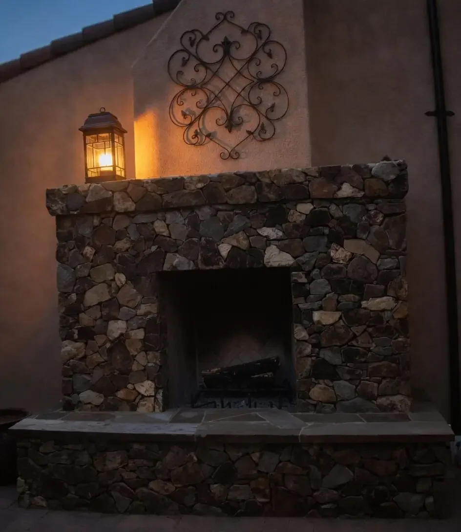 Exterior fireplace made of stone with a decorative wrought iron wall piece above it, and a lantern-style outdoor light on top emitting warm light, in evening setting.