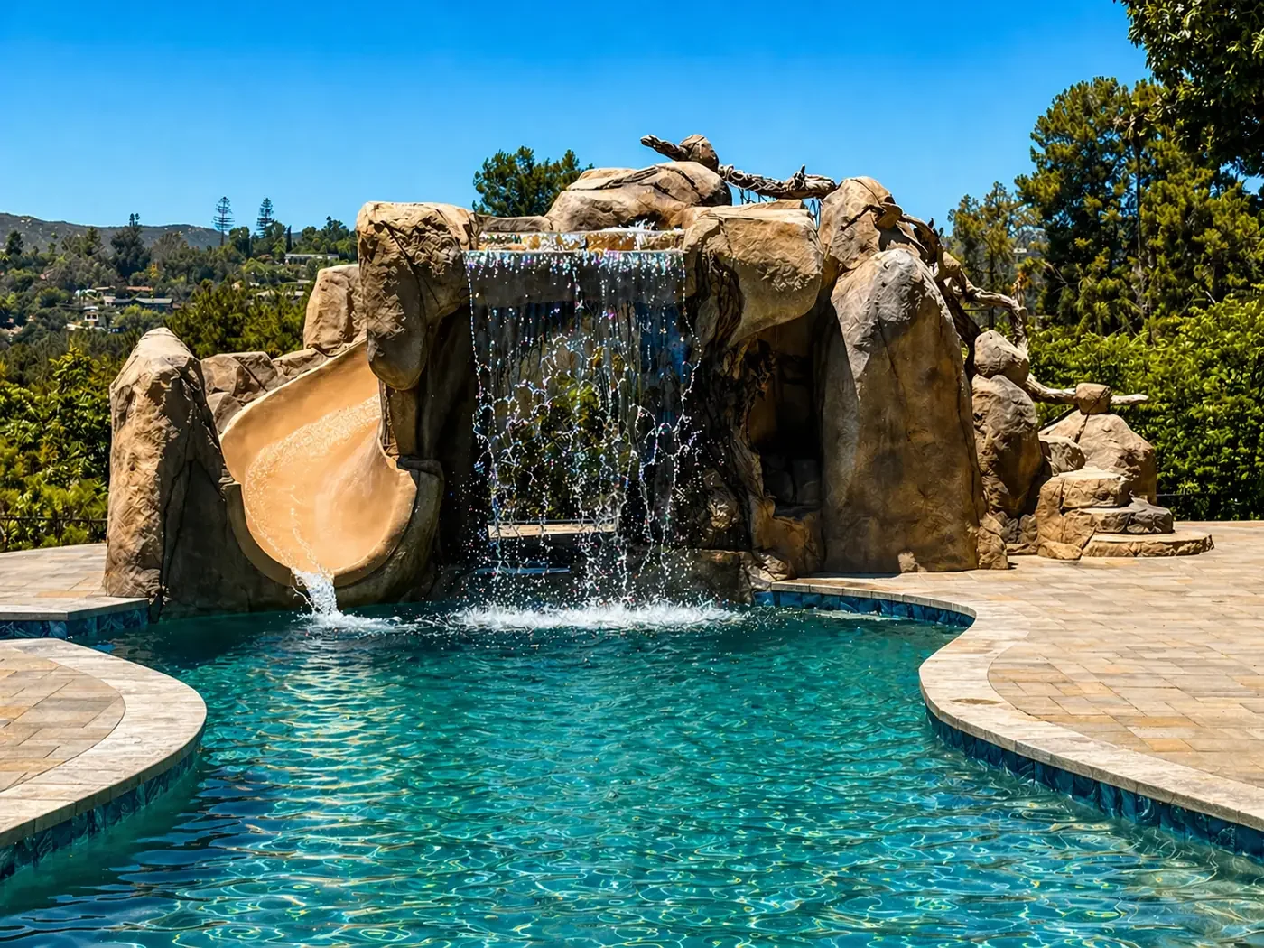 Luxury backyard pool with rock waterfall feature under sunny blue sky, surrounded by trees and hilly landscape.