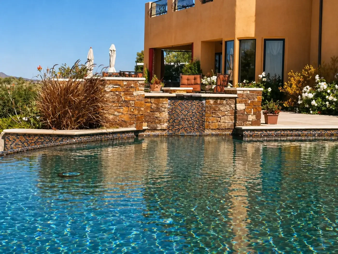 A modern backyard with a swimming pool featuring a waterfall, surrounded by a patio with outdoor furniture, potted plants, and flowering bushes, with a multi-story house in the background.