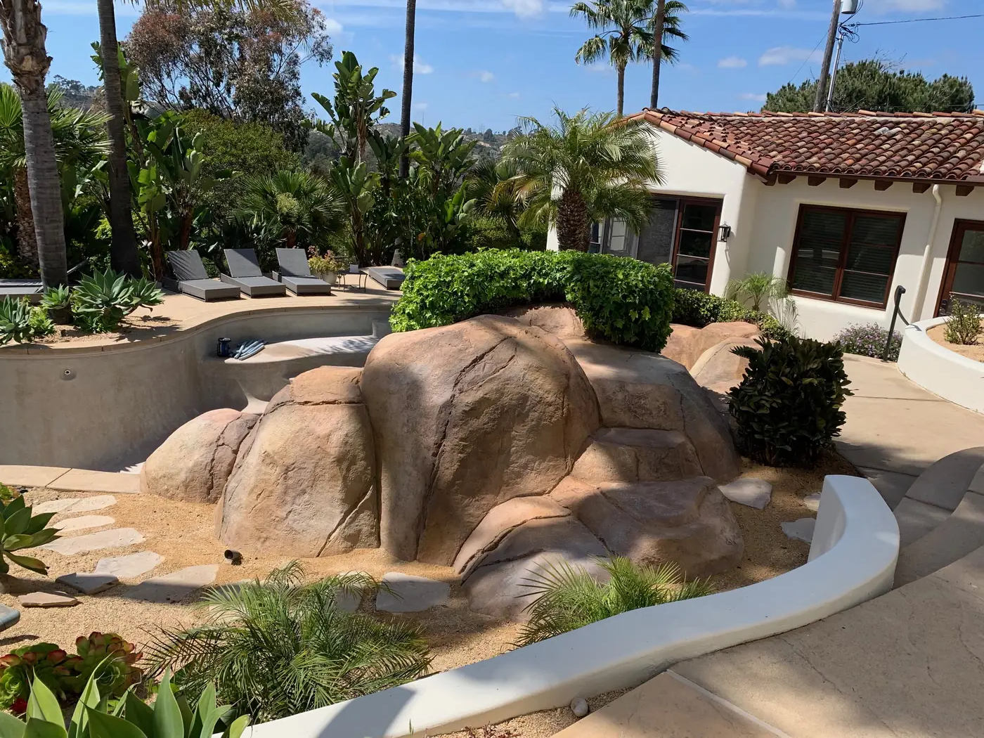 Backyard with large rocks, palm trees, lounge chairs, and a heated spa, with a white house in the background.