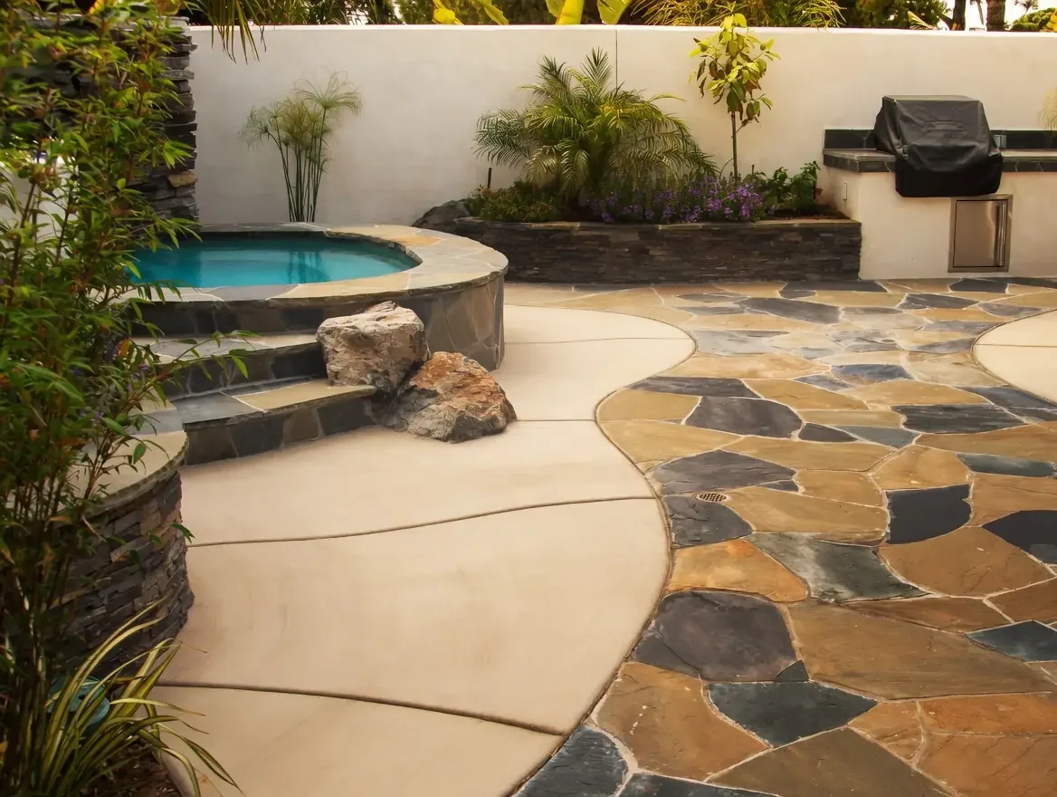 A backyard patio with a small hot tub, stone flooring, potted plants, and a built-in grill area.