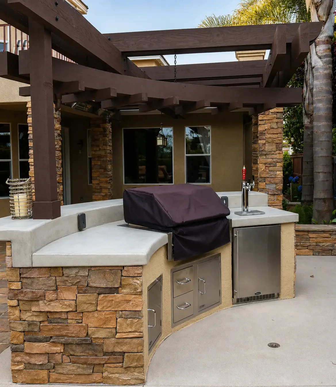 Outdoor kitchen with a built-in grill covered by a black protective cover, beige stone and concrete countertop, stainless steel cabinets, and a beverage tap, under a wooden pergola with brick columns, in a backyard with windows and palm trees.