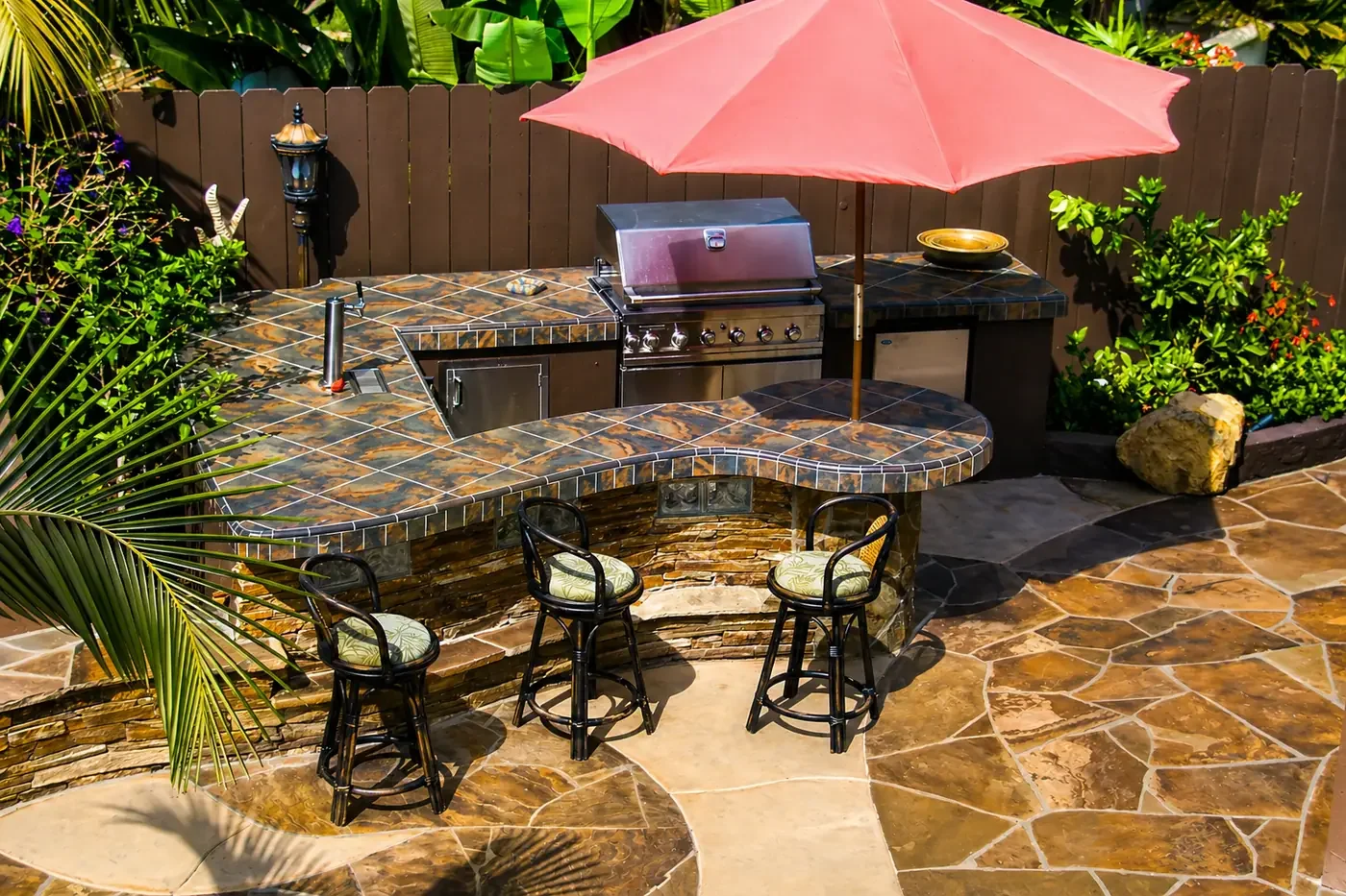 Outdoor kitchen with a tiled stone bar, a gas grill, a pink patio umbrella, three bar stools with patterned cushions, surrounded by lush green plants and rocks.