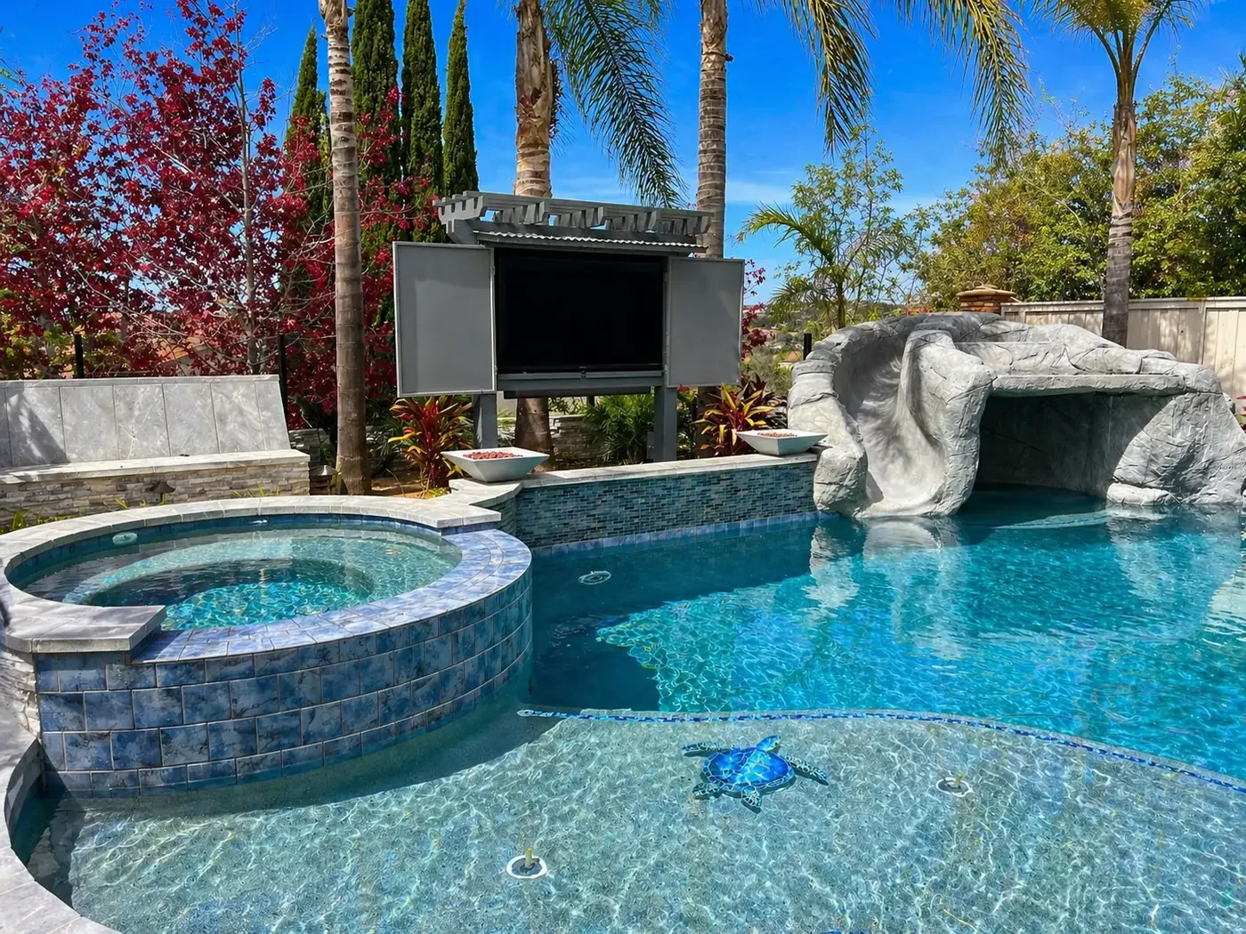 A backyard swimming pool with a hot tub, a water slide, surrounded by tropical plants and trees, with a clear blue sky overhead.