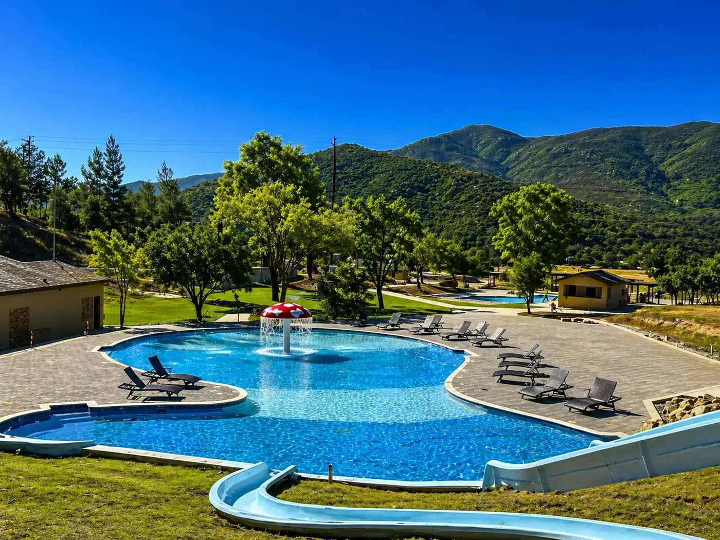 A backyard swimming pool with a mushroom water fountain, surrounded by lounge chairs and trees, with green mountains in the background under a clear blue sky.