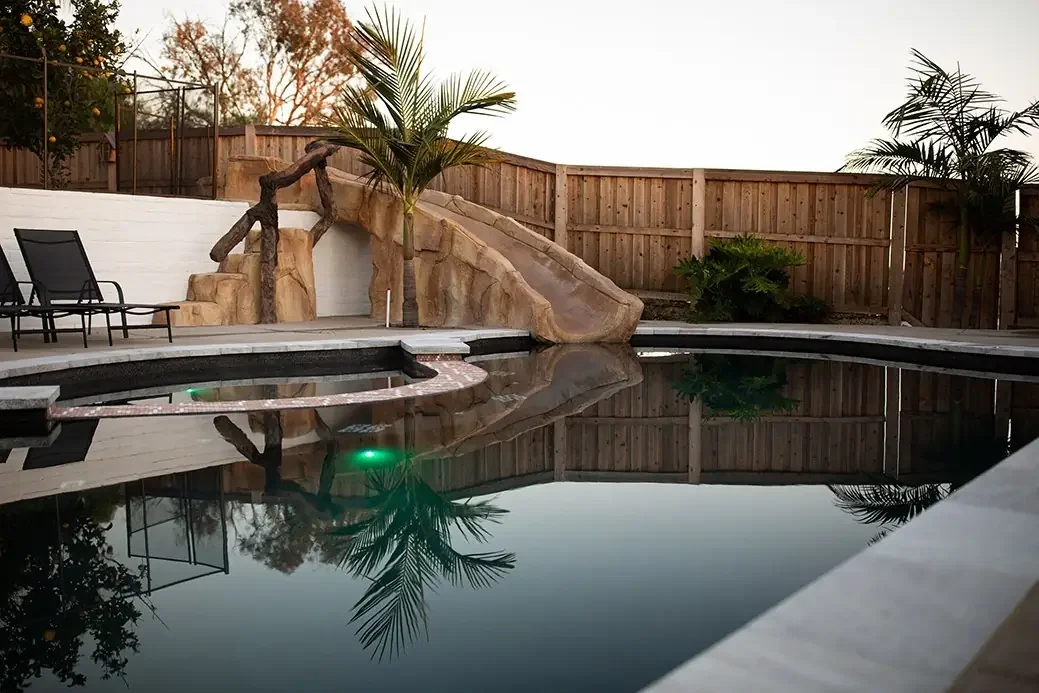 Backyard pool with a rock slide, palm trees, and lounge chairs, surrounded by a wooden fence at sunset.