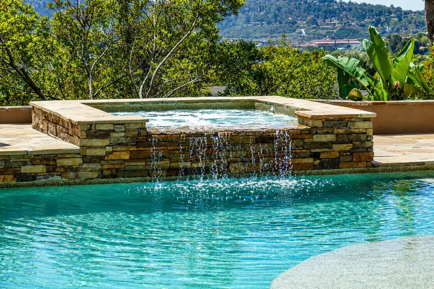 A backyard swimming pool with a stone water feature waterfall and lush greenery in the background.