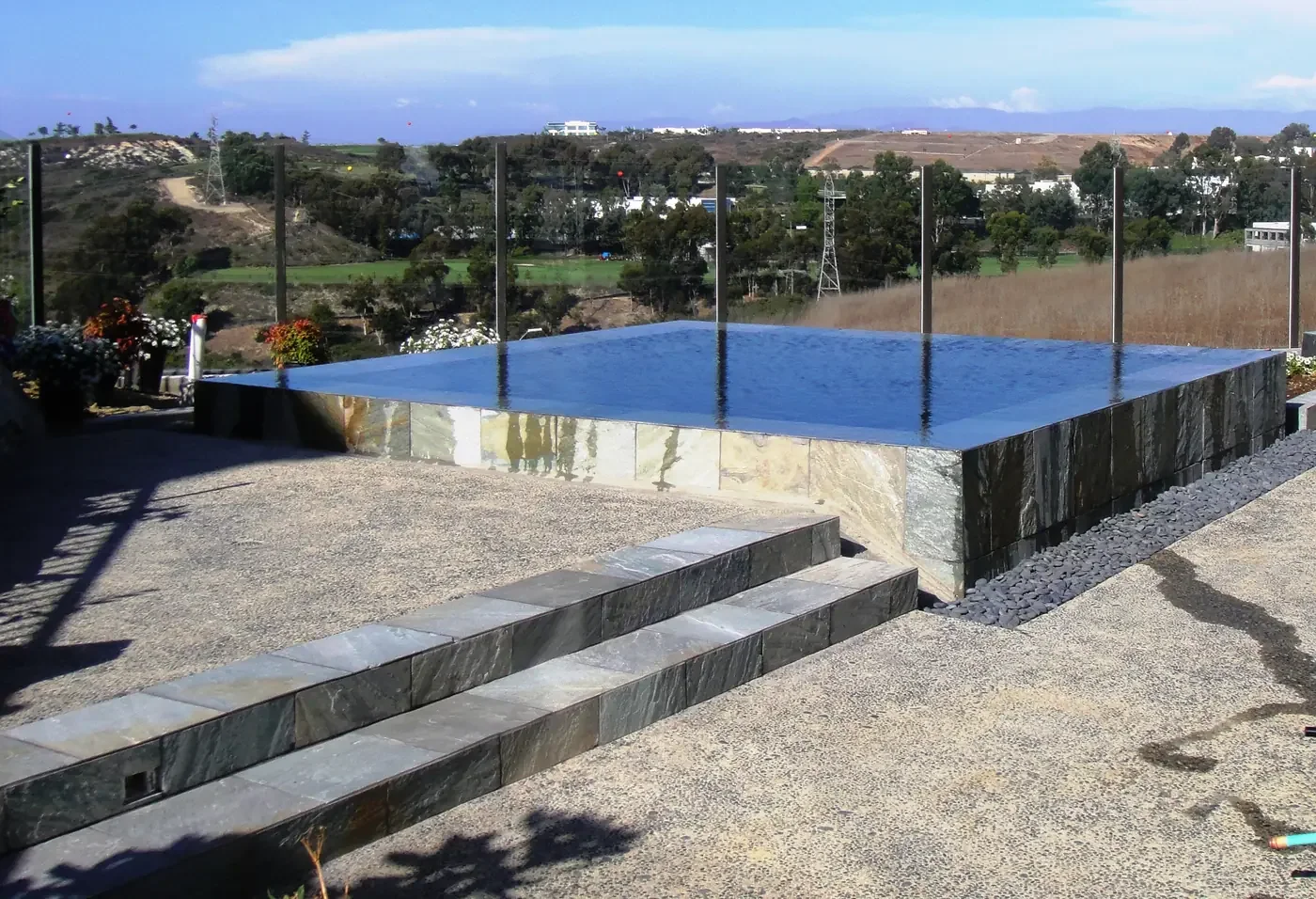 A modern outdoor infinity pool on a patio with stone steps and glass railing, overlooking a hilly landscape with trees and open fields.