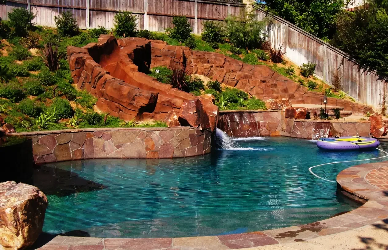 Backyard swimming pool with rock waterfall feature and landscaped plants, enclosed by a wooden fence.