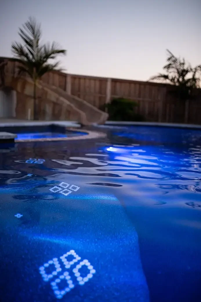 A backyard swimming pool at dusk with a wooden fence, palm trees, and an inflatable slide, illuminated by blue pool lights.