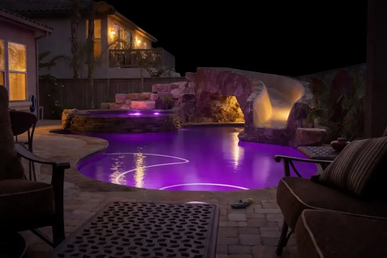 Nighttime scene of a backyard with a lit swimming pool illuminated with purple and yellow lights, surrounded by stonework and outdoor furniture.