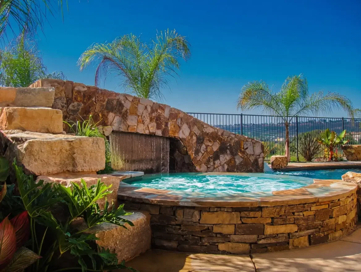 Luxury outdoor stone hot tub with water feature, surrounded by lush green plants and palm trees under a blue sky.