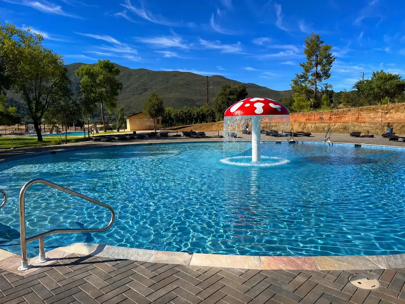An outdoor swimming pool with a mushroom-shaped water fountain in the center, surrounded by trees, lounge chairs, and mountains in the background under a blue sky.