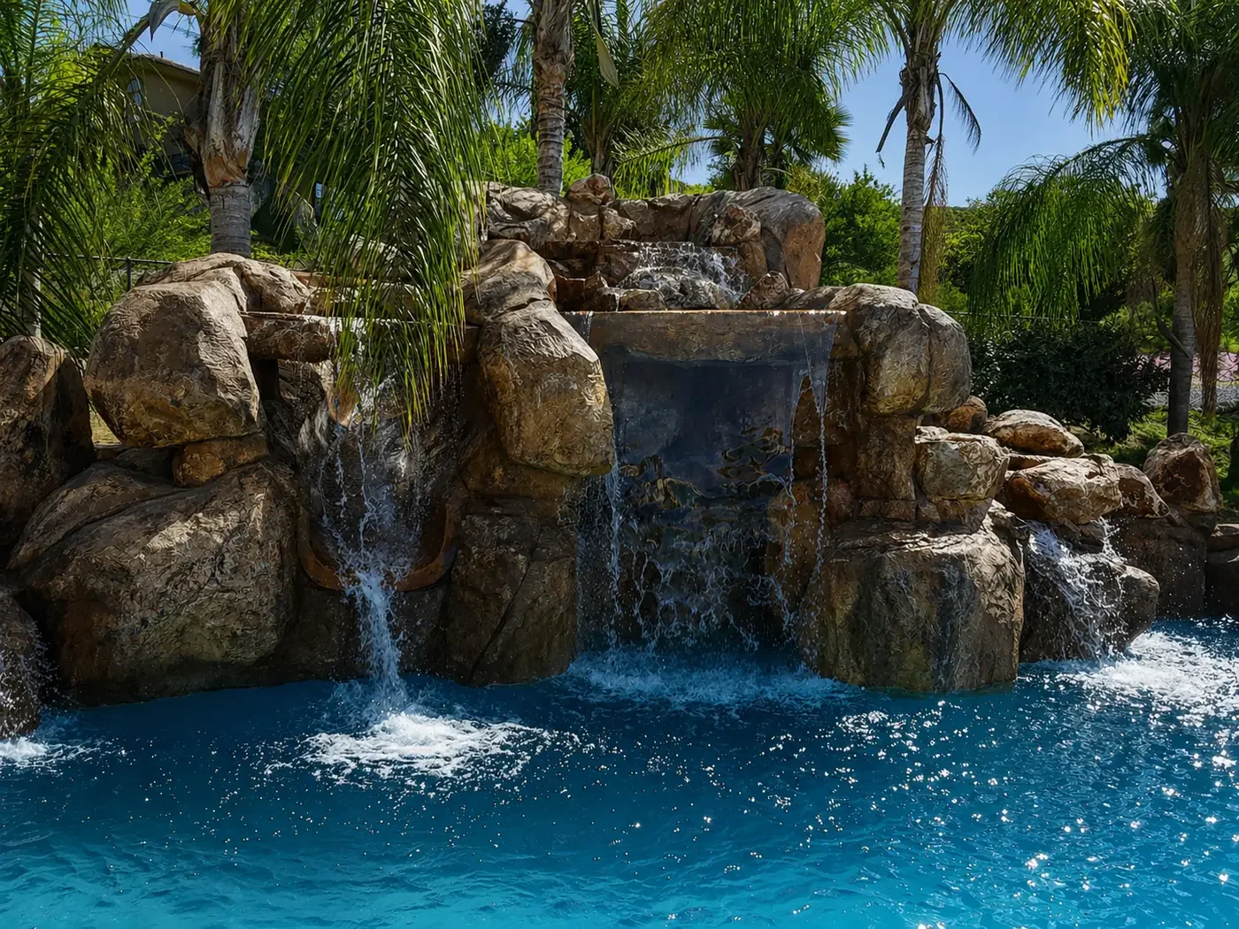 A man-made waterfall with water cascading over large rocks into a blue pool, surrounded by palm trees and lush greenery.