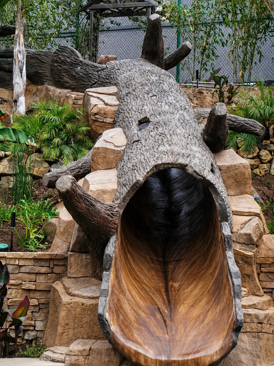 A large wooden animal-shaped slide in a botanical garden setting with rocks and lush green plants surrounding it.