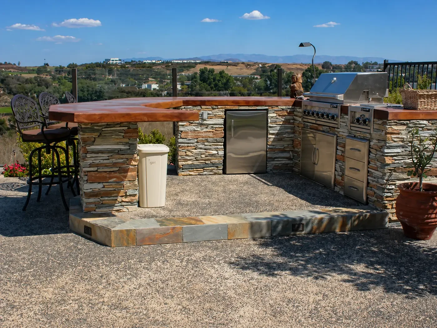 Outdoor kitchen and bar with stone and wood construction, metal grill, small refrigerator, and bar stools, overlooking a scenic landscape under a blue sky.