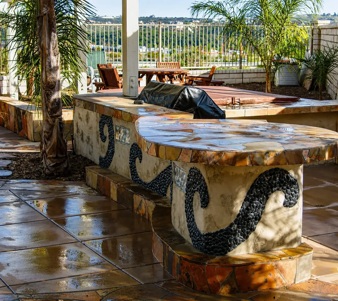 Outdoor patio with tiled tables and chairs, palm trees, and decorative stonework, overlooking a distant landscape.