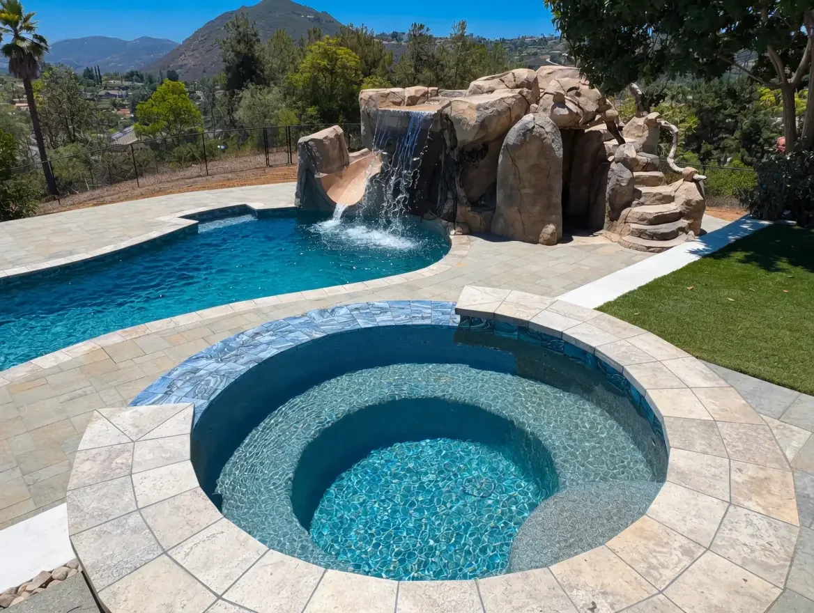 Swimming pool and hot tub in backyard with waterfall feature and rocky structure, surrounded by paved patio and greenery, mountainous landscape in the background.