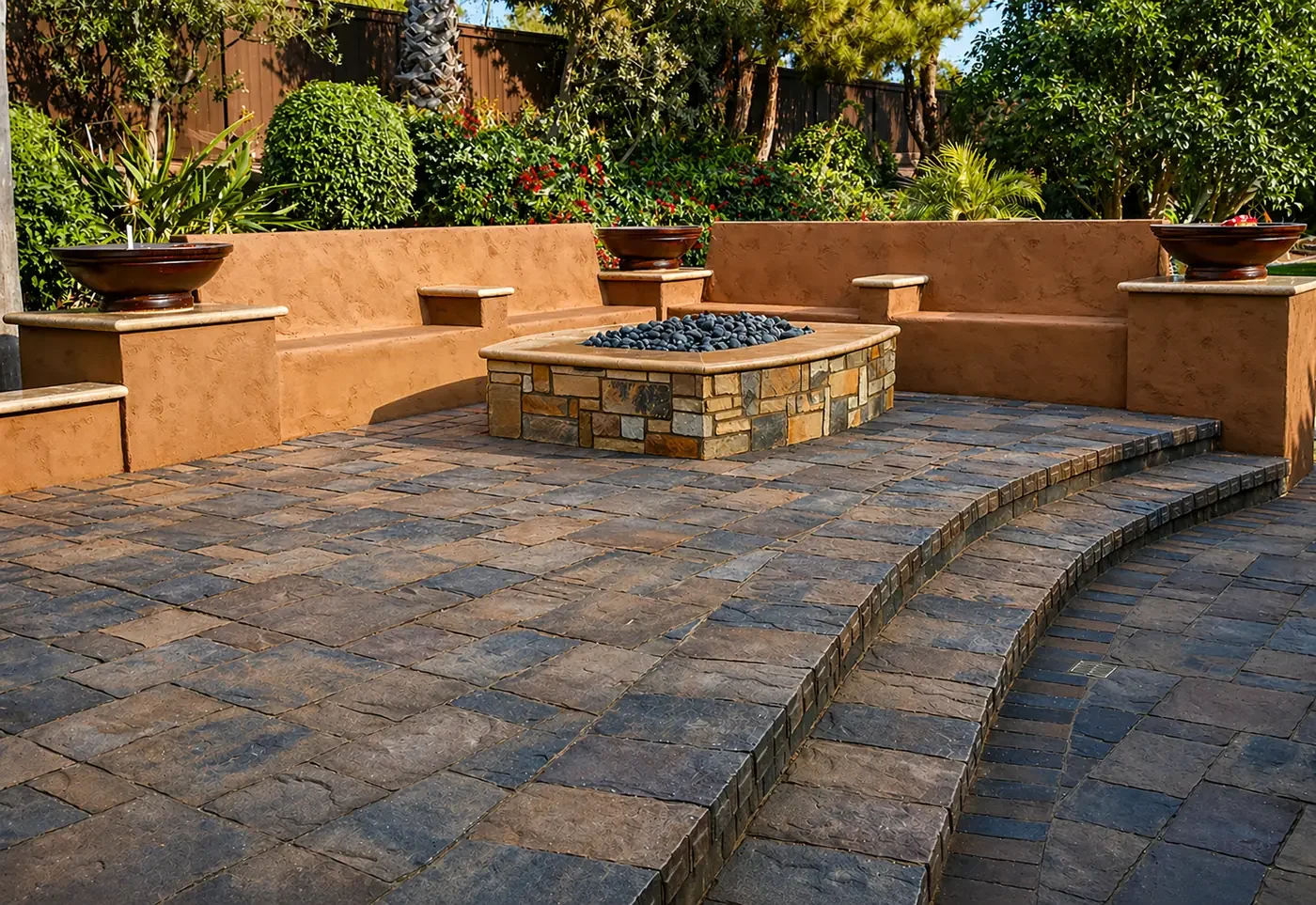 A backyard patio with curved stone stairs leading to a firepit surrounded by a low stucco wall. The firepit is filled with black stones and there are built-in seats with large flower pots on ledges above the seats. Green shrubs and trees are in the b