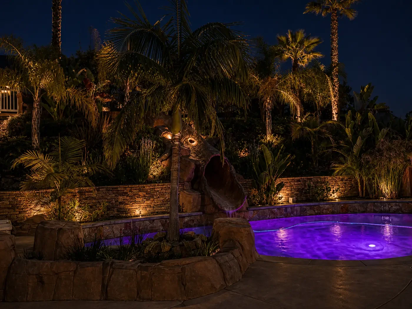 A night view of a backyard pool area illuminated with purple lighting, surrounded by lush tropical plants and palm trees, with a stone water slide and decorative lighting on the trees and plants.
