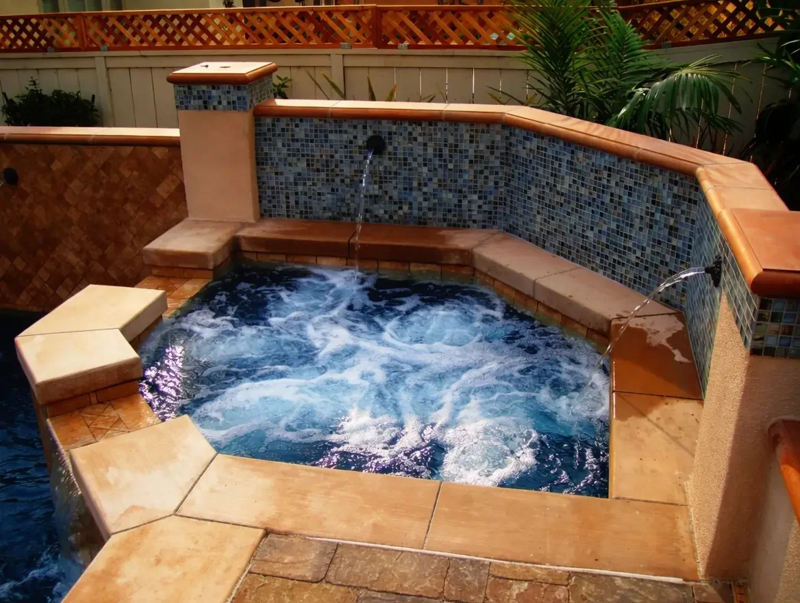 A small hot tub with bubbling water, built with stone and tile, surrounded by a fence and tropical plants.