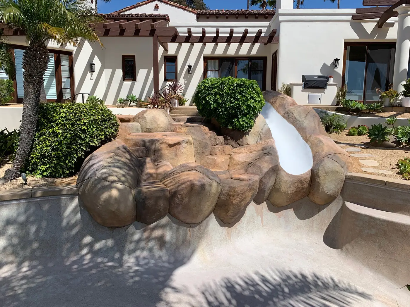 A backyard with a small waterfall and slide built into rocks, surrounded by green bushes and plants, with a white house in the background.