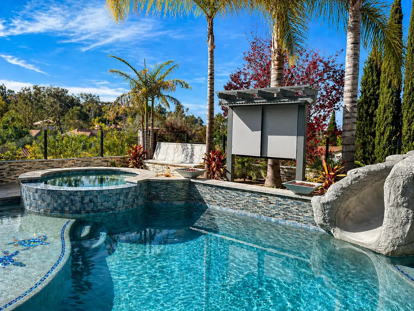A backyard swimming pool with a hot tub, surrounded by tropical plants and tall palm trees, under a bright blue sky with some clouds.