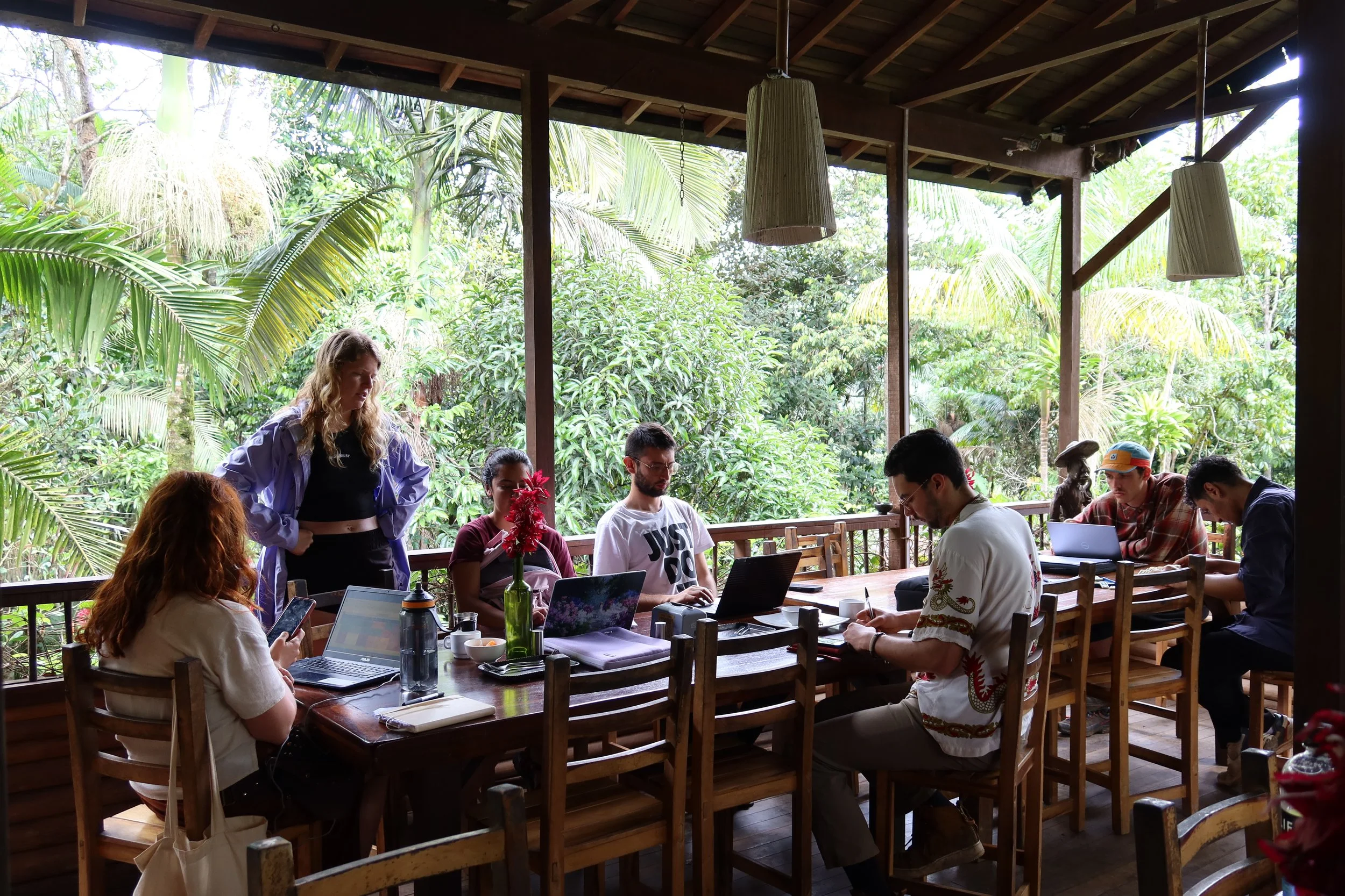 People working on laptops and phones around a wooden table on a covered patio with lush green trees in the background.