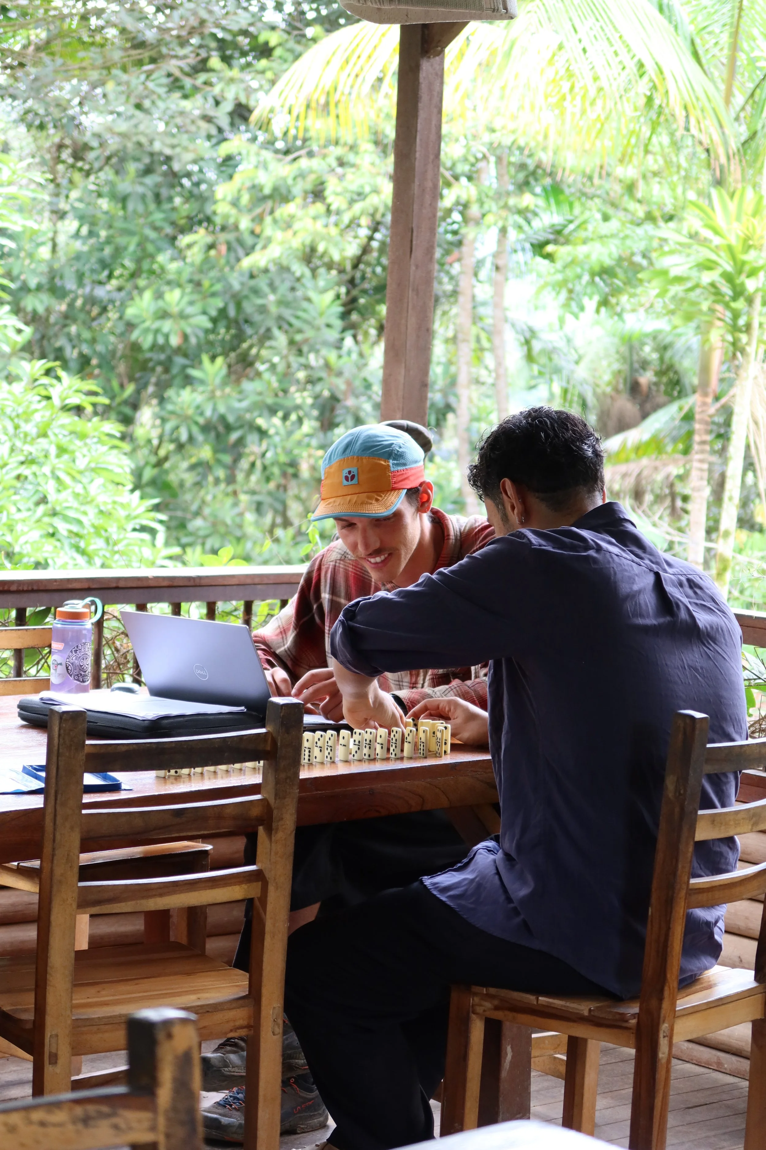 Two young men are playing dominoes at a wooden table outdoors, surrounded by lush greenery.