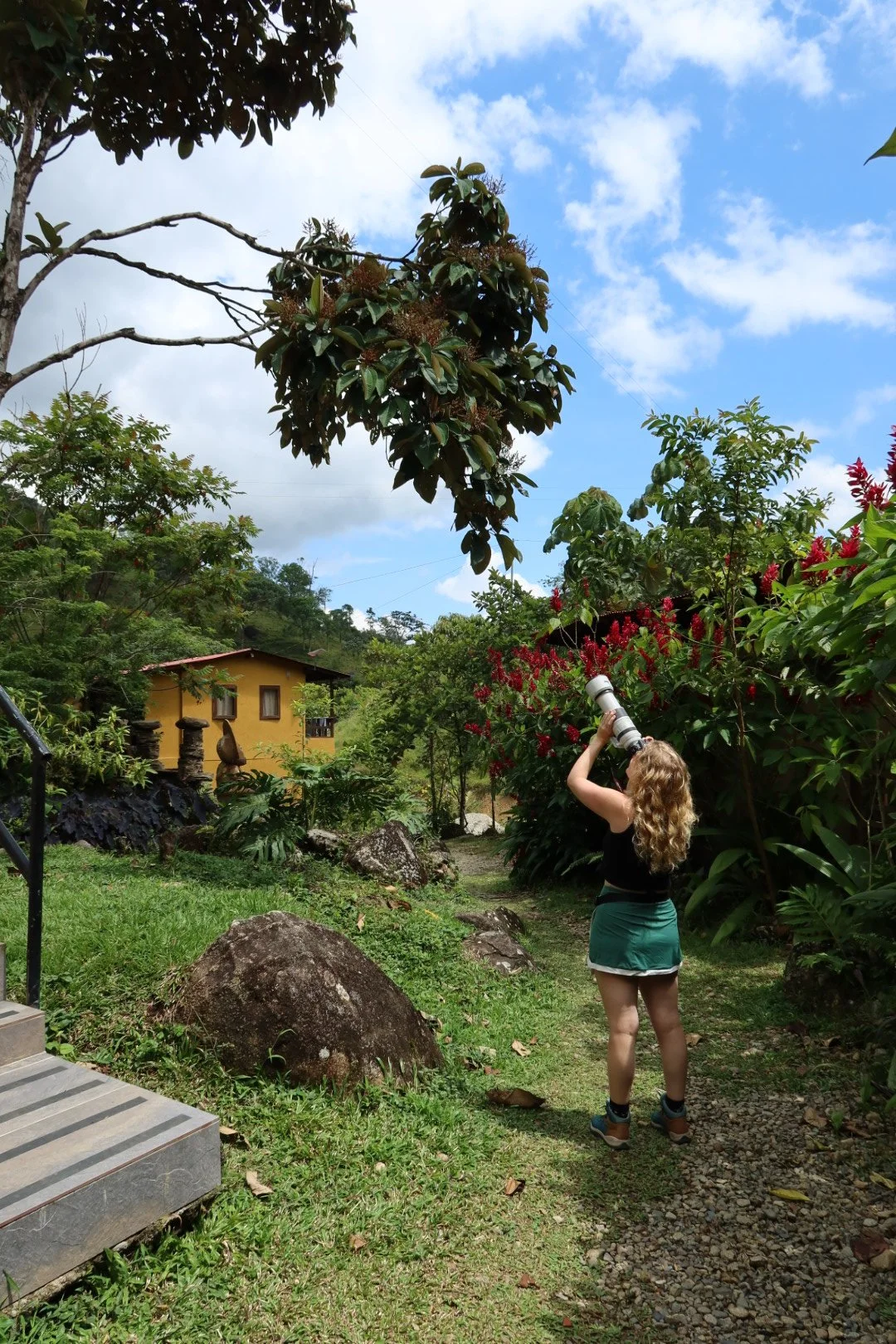 A woman with curly blonde hair takes a photo with a camera lens in a lush garden with green trees, shrubs, and a small yellow house against a partly cloudy sky.