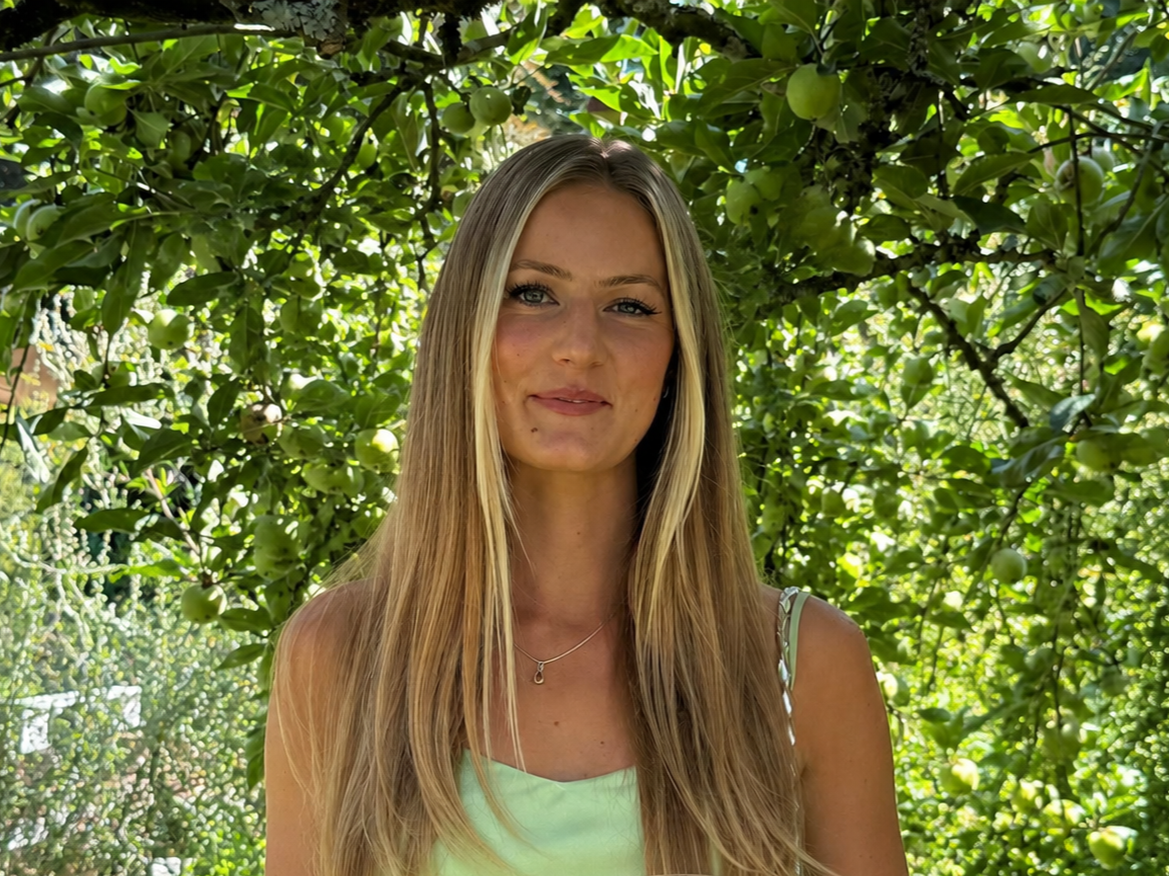 A young woman with long blonde hair smiling outdoors in front of an apple tree with green apples and bright green leaves.