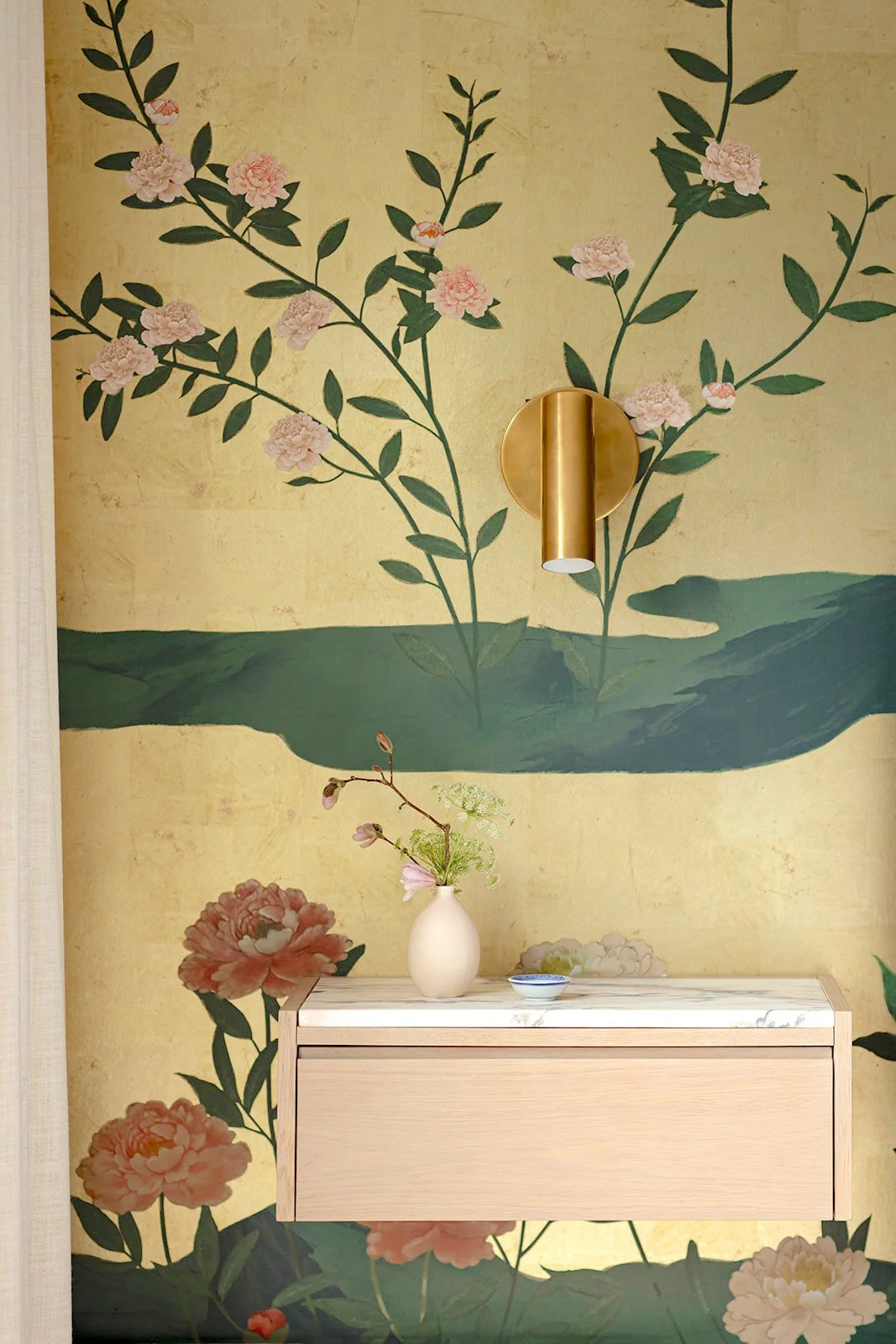 A wall with floral mural, a gold wall sconce, a white vase with flowers on a wooden shelf, and a small dish.