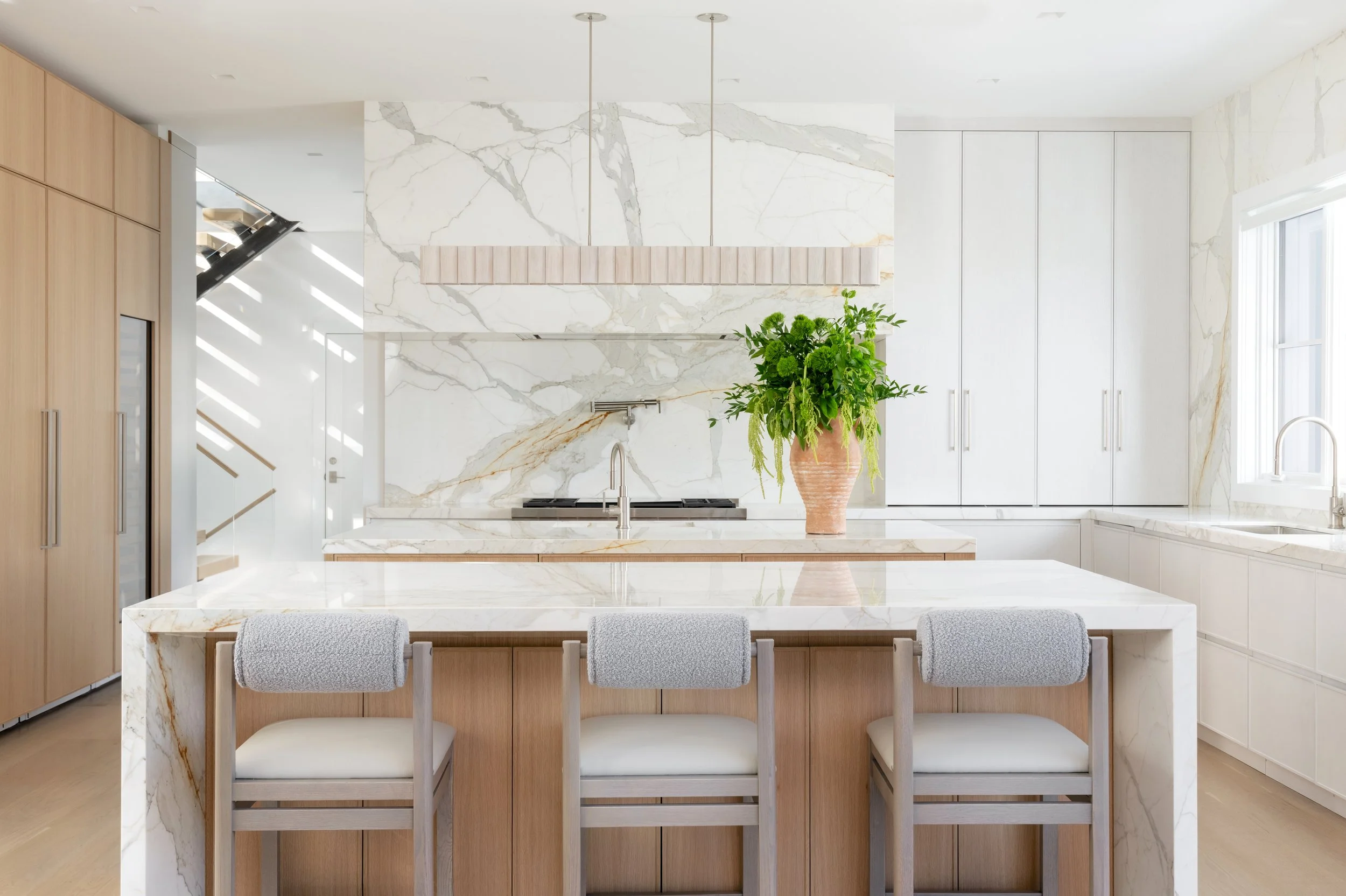 Modern white kitchen with marble countertops and backsplash, wooden cabinetry, a large island with three bar stools, a plant in a peach-colored vase, and a window on the right side.
