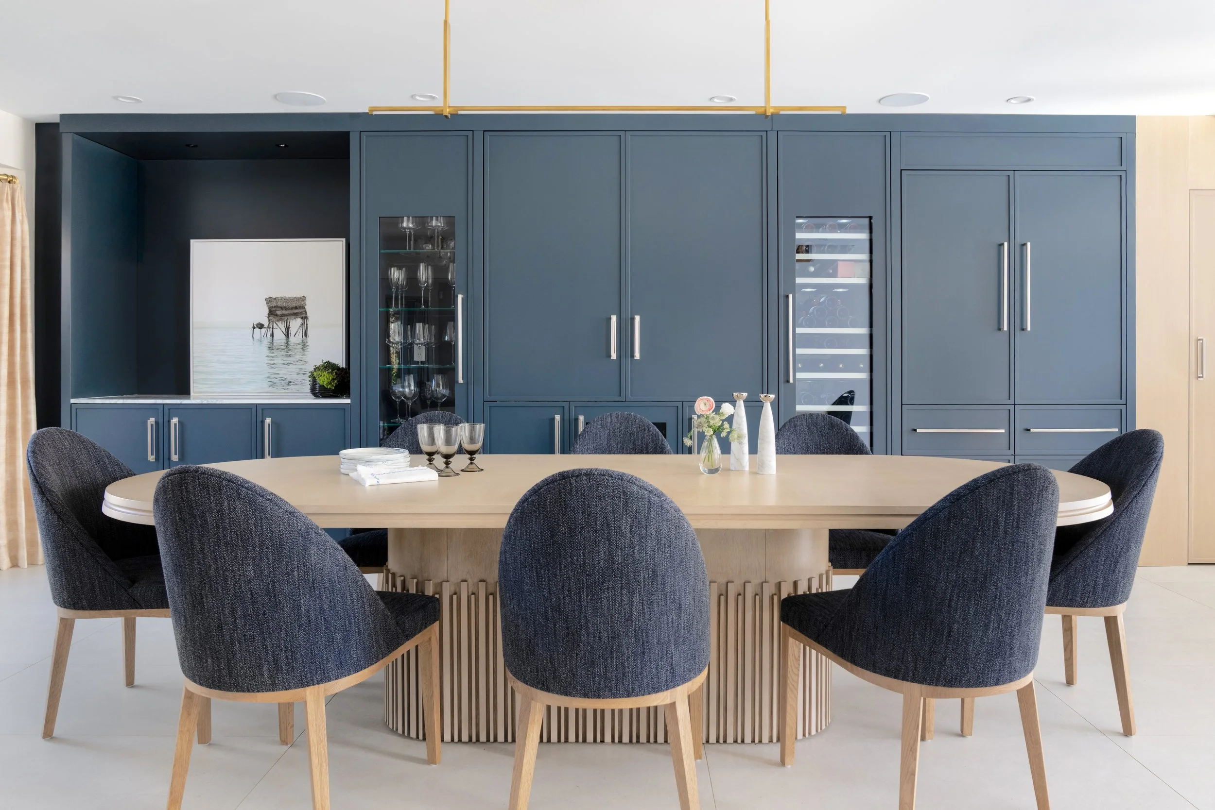 Modern dining room with a large wooden table and six dark upholstered chairs, navy built-in cabinets with glassware and wine storage, artwork, and decorative vases in the center.